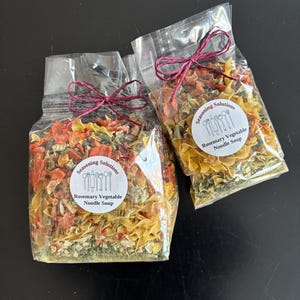 May include: Two clear plastic bags filled with Rosemary Vegetable Noodle Soup. The bags are tied with a burgundy ribbon. Each bag has a white label with the text "Seasoning Solutions" and "Rosemary Vegetable Noodle Soup". The soup mix contains pasta, vegetables, and herbs.