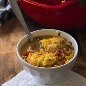 May include: A white bowl filled with a creamy soup topped with corn kernels, diced tomatoes, and golden brown biscuits. The soup is garnished with fresh herbs.