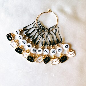 May include: Ten black and white cat stitch markers with gold rings and black string. The markers are numbered 0 through 9.