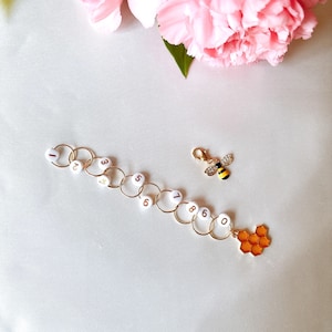 May include: A gold-coloured stitch marker set with white numbered beads and a bee charm. The set includes a honeycomb charm and a bee charm with black and yellow stripes. Pink flowers are in the background.