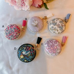 May include: Five round fabric-covered measuring tapes labelled A through E. A features pink flowers, B has white flowers on lavender, C shows blue floral patterns, D displays a bird and strawberry motif, and E has purple flowers. Each has a tassel.