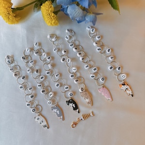 May include: A selection of cat-themed keyrings. Each keyring has a chain of white beads with numbers and a small cat charm. The cat charms are in various colours and designs. A gold-coloured fishbone charm is also included.