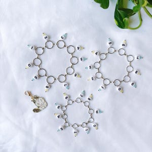 Peut inclure: Trois bracelets en chaîne argent avec des perles blanches numérotées de 0 à 9. Les perles sont rehaussées d'accents bleu clair et jaune.