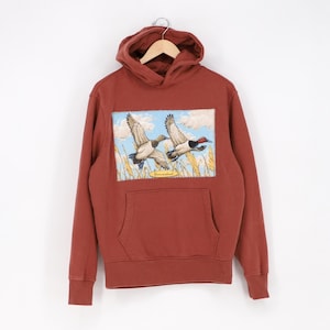May include: A rust-colored hooded sweatshirt with a front pocket and a graphic of two ducks in flight over a field of wheat. The ducks are depicted with detailed feathers and vibrant colors against a blue sky.