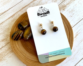 Puede incluir: Un par de pendientes de oro con cuentas de ojo de tigre. Los pendientes se muestran en una tarjeta blanca con el texto "NORTHERN solstice" y "crafted with magick + intention northern-solstice.ca".