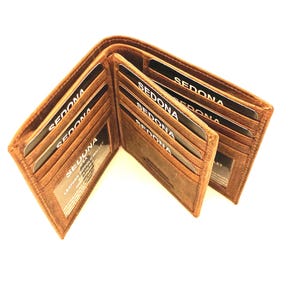 May include: A tan leather bifold wallet featuring multiple interior card slots and a clear identification window. Several black cards with the white text SEDONA are inserted into the slots. The wallet has visible stitching along the edges.
