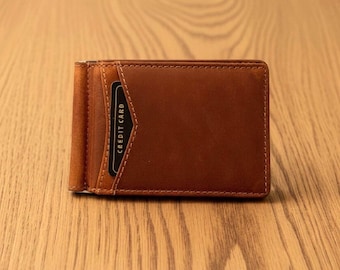 Könnte beinhalten: Eine braune Ledergeldbörse mit Geldklammer, flachem Profil, sichtbaren Nähten an den Kanten und einem äußeren Kartenfach, das eine schwarze Karte mit der Aufschrift CREDIT CARD enthält. Oben steht der Text RFID Protected.