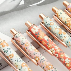 May include: Close-up of several floral-patterned pens with gold-colored accents. The pens feature a variety of colors, including red, pink, yellow, and blue, with a floral design. The pens are arranged in a row, showcasing their decorative patterns.