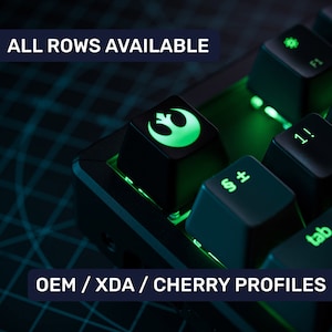 Può includere: Una tastiera meccanica nera con retroilluminazione verde. La tastiera ha un cappuccio per tasti con un simbolo verde e bianco della Ribellione di Star Wars. Il testo "ALL ROWS AVAILABLE" e "OEM / XDA / CHERRY PROFILES" è visibile sulla tastiera.