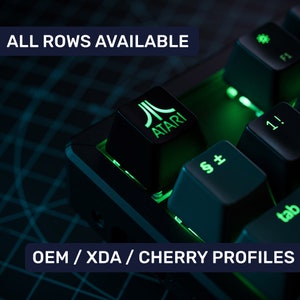 May include: A black mechanical keyboard with green backlighting. The keyboard has a keycap with the Atari logo and the text "ATARI". The keyboard also has keys labeled "§±", "1!", "tab", and "F1". The text "ALL ROWS AVAILABLE" and "OEM/XDA/CHERRY PROFILES" is visible in the image.