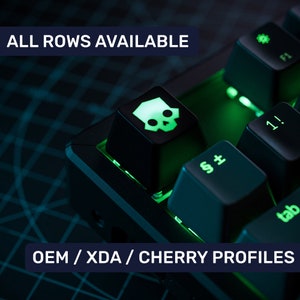 May include: A black mechanical keyboard with green backlighting. The keyboard has a skull keycap on the space bar. The text "ALL ROWS AVAILABLE" and "OEM/XDA/CHERRY PROFILES" is visible on the keyboard.