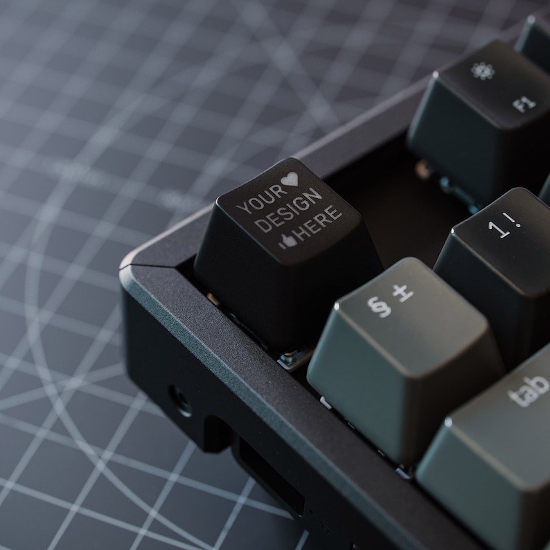 Peut inclure: Un clavier m&eacute;canique noir avec une touche personnalis&eacute;e qui dit "YOUR DESIGN HERE" avec un c&oelig;ur et un pouce en l'air. Le clavier a un cadre noir et des touches grises.