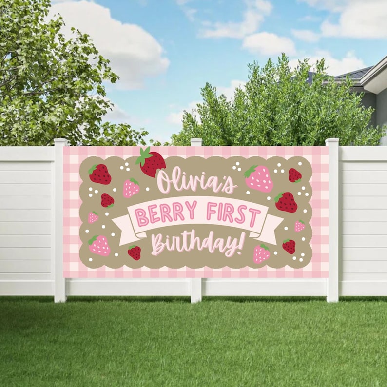 May include: A pink and brown banner with the text "Olivia's BERRY FIRST Birthday!" and strawberry illustrations. The banner has a pink gingham background and is displayed on a white fence.
