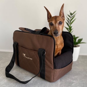 May include: A brown and black pet carrier with a black strap and the word "HAUSTIER" embroidered on the front. A small brown dog is sitting inside the carrier.