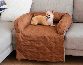 May include: A small, light brown chihuahua dog is lying on a brown quilted dog bed on a gray couch. The dog bed has a brown quilted cover and a brown fabric border. The couch has a gray fabric cover and a white pillow.