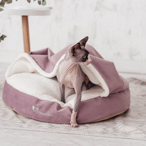 May include: A hairless cat is curled up in a plush, mauve-coloured pet bed with a white fleece lining. The bed has a round shape and is designed with a raised edge. The cat is looking to the right of the image, and the bed has the word "Haustier" embroidered on the bottom.