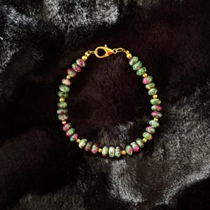 May include: A beaded bracelet featuring green and red gemstones with gold-colored spacers and a clasp. The bracelet is made of small, round beads, and the clasp is a lobster claw style. The bracelet is displayed on a black, textured surface.