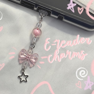 May include: A mobile device accessory charm featuring a silver-toned metal star, a translucent pink bow-shaped bead, and a round pink bead attached to a clear plastic connector. The text E-reader charms is written in a cursive white font.