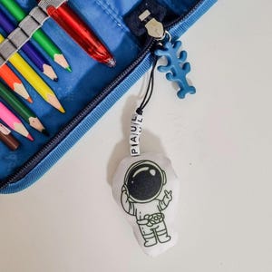 Könnte beinhalten: Ein blaues Federmäppchen aus Stoff mit Buntstiften und einem roten Stift, an dessen Reißverschluss ein weißer Plüsch-Astronauten-Schlüsselanhänger befestigt ist. Der Anhänger hat eine schwarze Schnur mit weißen Buchstabenperlen, die PAUL bilden.