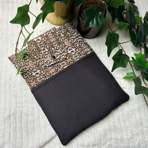 Op de afbeelding: Een zwarte en luipaardprint stoffen hoes of etui. De bovenklep heeft een luipaardprintpatroon met een zwarte knoopsluiting. De onderste helft is effen zwart. De hoes is waarschijnlijk voor een tablet of e-reader.