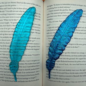 Op de afbeelding: Twee turquoise blauwe glitter veren bladwijzers met een citaat uit een boek over herinneren en vergeten. Het citaat luidt: "Het doet pijn om te herinneren; het doet pijn om te vergeten. En nu is alles hier dood."