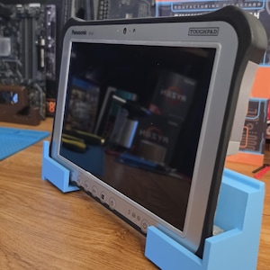 Op de afbeelding: Een Panasonic Toughpad tablet met een zwart en zilverkleurig frame, voorzien van een zwart scherm en fysieke knoppen aan de onderrand, stevig geplaatst in een lichtblauwe, op maat gemaakte kunststof standaard op een houten ondergrond.