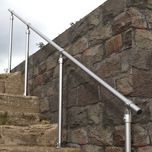 May include: A silver handrail is mounted to a stone wall and extends down a set of stone steps. The handrail is made of metal pipes and is secured to the wall with brackets.