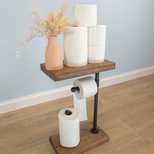 May include: A rustic, industrial-style toilet paper holder. It features a dark wooden base and shelf, connected by black metal pipes. The shelf holds several rolls of white toilet paper and a peach-colored vase with dried flowers. Another roll of toilet paper is on the holder.