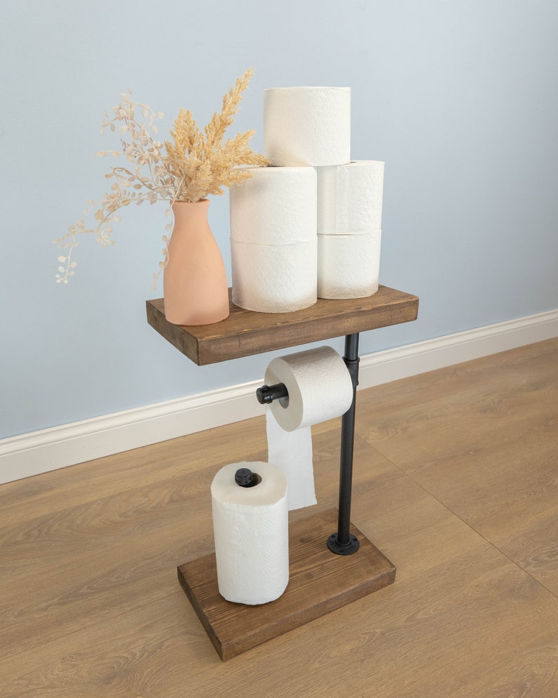 May include: A rustic, industrial-style toilet paper holder. It features a dark wooden base and shelf, connected by black metal pipes. The shelf holds several rolls of white toilet paper and a peach-colored vase with dried flowers. Another roll of toilet paper is on the holder.