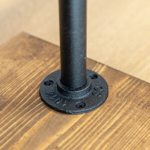 May include: Close-up of a black metal pipe attached to a wooden surface. The pipe is secured with a round flange and screws. The wood has a natural grain and a warm, brown color.