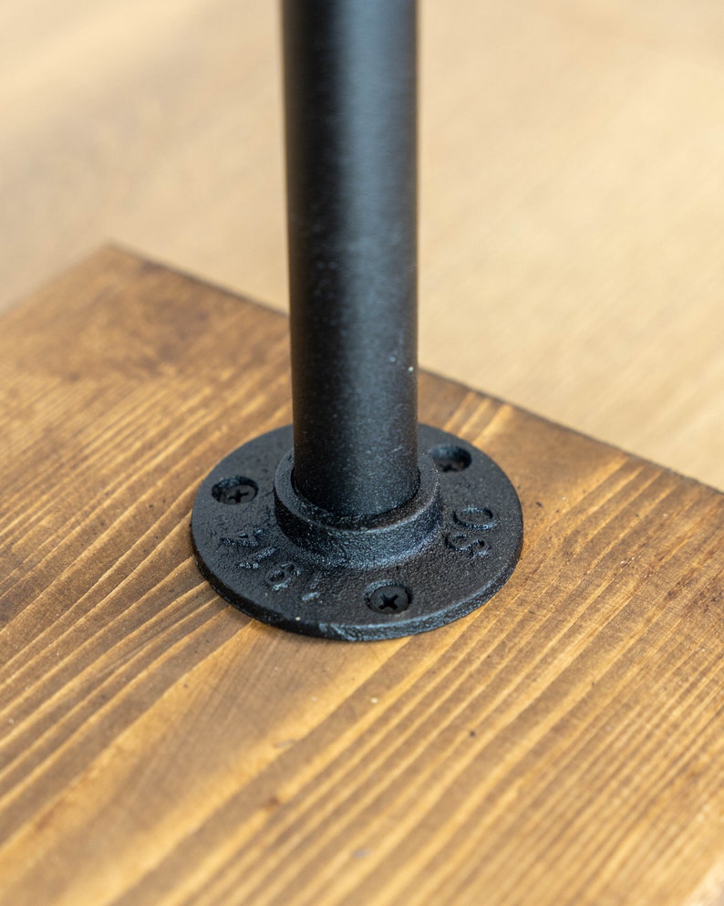 May include: Close-up of a black metal pipe attached to a wooden surface. The pipe is secured with a round flange and screws. The wood has a natural grain and a warm, brown color.