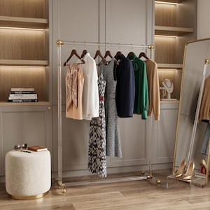 Puede incluir: Un perchero de ropa de acrílico transparente con detalles dorados sostiene varias prendas, incluyendo vestidos, una blazer y suéteres. Un espejo de cuerpo entero y un pequeño otomán también están en la habitación. La habitación tiene una paleta de colores neutros.