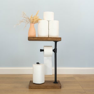 May include: A rustic, industrial-style toilet paper holder. It features a dark wooden base and shelf, with black metal piping. The holder displays several rolls of white toilet paper and a small vase with dried flowers. The design is functional and decorative.