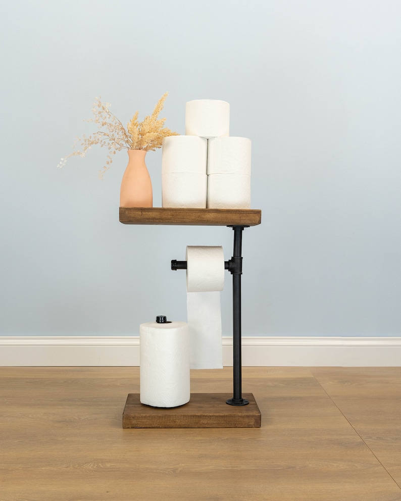 May include: A rustic, industrial-style toilet paper holder. It features a dark wooden base and shelf, with black metal piping. The holder displays several rolls of white toilet paper and a small vase with dried flowers. The design is functional and decorative.