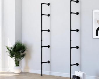 May include: Two black metal pipe shelves mounted on a white wall. Each shelf has five circular platforms. A potted plant and shopping bags are in the foreground. The shelves are a modern, industrial design.