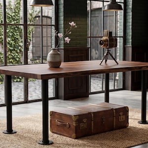 Könnte beinhalten: Ein rechteckiger Holzesstisch mit natürlicher Baumkante, getragen von vier zylindrischen schwarzen Metallbeinen. Eine Keramikvase mit einem Blumenstiel und eine Vintage-Kamera auf einem Stativ stehen auf der Tischplatte, während ein brauner Koffer darunter steht.