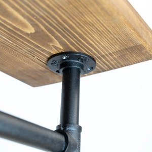 May include: Close-up of a wooden table top with a dark stain, supported by black metal pipes and fittings. The wood grain is visible. The table is designed with an industrial aesthetic.
