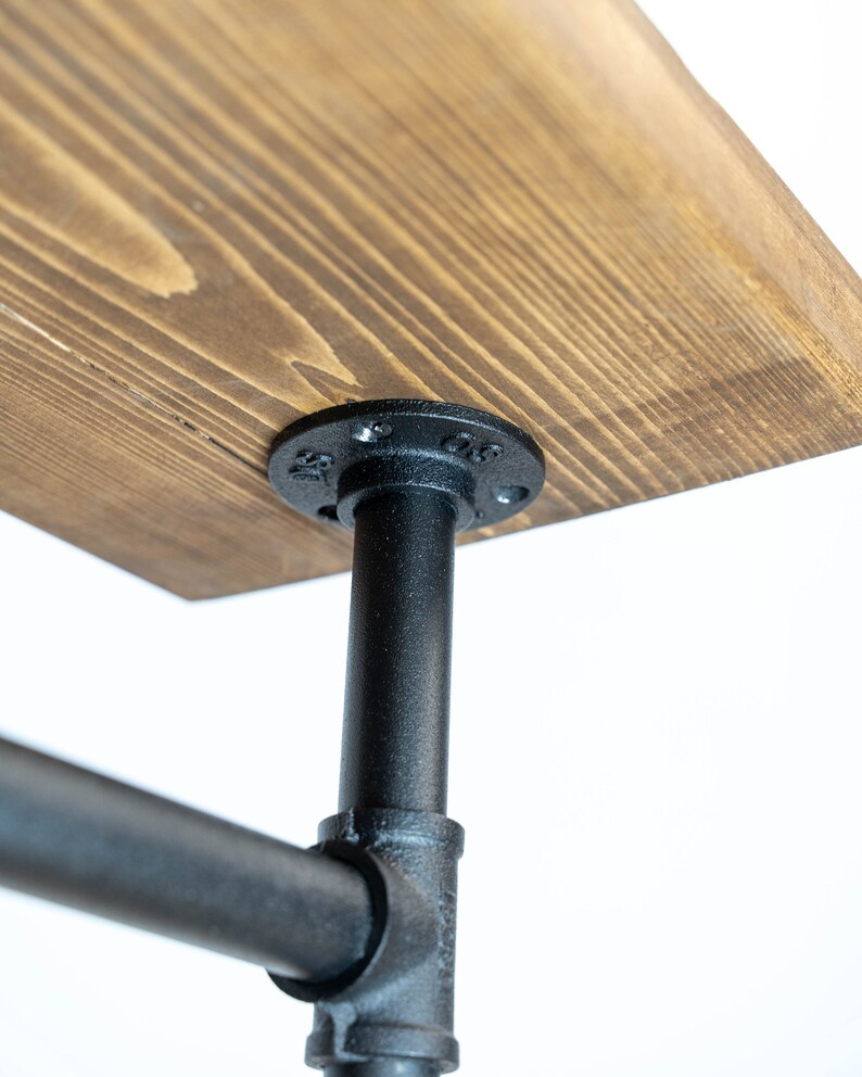 May include: Close-up of a wooden table top with a dark stain, supported by black metal pipes and fittings. The wood grain is visible. The table is designed with an industrial aesthetic.
