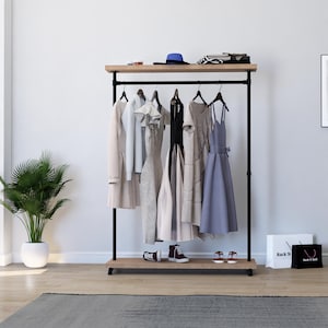 Pode incluir: Um cabide de metal preto com uma prateleira superior e inferior de madeira. O cabide tem uma barra de pendurar única e está segurando uma variedade de roupas, incluindo vestidos, camisas e uma jaqueta. O cabide está em pé em um piso de madeira com um tapete na frente.