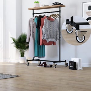 Pode incluir: Um cabideiro de metal preto com uma prateleira de madeira e roupas penduradas. O cabideiro tem uma prateleira superior, uma barra para pendurar e rodas. As roupas incluem um casaco cinza claro, um vestido turquesa e um suéter cinza.