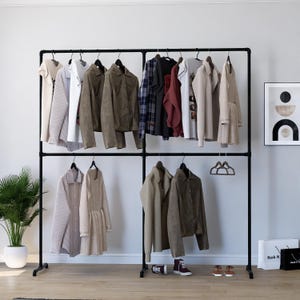 May include: A black metal clothing rack with two tiers of hanging rails. The rack is filled with various clothing items, including jackets, shirts, and dresses. There are also shoes and a bag on the floor in front of the rack.