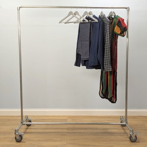 Pode incluir: Um cabide de metal prateado com rodas. O cabide tem uma barra horizontal com cinco cabides segurando roupas. As roupas incluem uma camisa azul, uma camisa xadrez, uma calça e um vestido de crochê colorido.