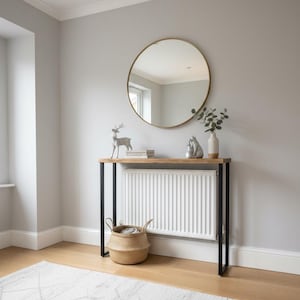 Puede incluir: Una habitación con un espejo redondo con marco dorado, una consola de madera con patas de metal negro y una cesta tejida. La mesa está decorada con una figura de ciervo, libros y un jarrón con vegetación. Una alfombra blanca con un patrón geométrico está en el suelo.