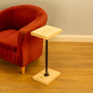Puede incluir: Una mesa auxiliar de madera con una pata de tubo de metal negro. La mesa tiene una tapa rectangular y una base cuadrada. La mesa está al lado de un sillón rojo.