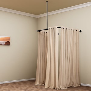 Puede incluir: Un sistema de barra de cortina de metal negro con cortinas beige colgadas. La barra est&aacute; unida al techo y se extiende para crear un recinto cuadrado.