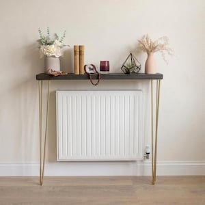 May include: A dark wood console table with gold hairpin legs. The table is decorated with a vase of flowers, books, a small terrarium, a vase of pampas grass, and other decorative items. The table is placed in front of a white radiator.