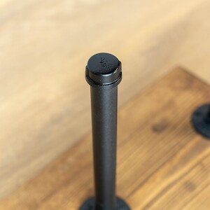 May include: Close-up of a black metal pipe with a cap, attached to a wooden surface. The pipe is cylindrical and has a threaded cap. The wooden surface is stained a warm brown color. The pipe is part of a larger industrial design.