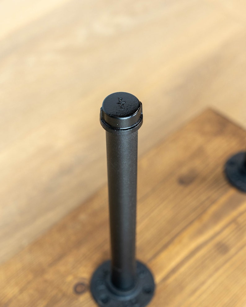 May include: Close-up of a black metal pipe with a cap, attached to a wooden surface. The pipe is cylindrical and has a threaded cap. The wooden surface is stained a warm brown color. The pipe is part of a larger industrial design.