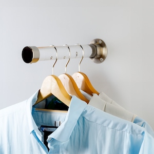 May include: Three wooden hangers with light blue shirts hanging on a silver metal rod mounted to a white wall.