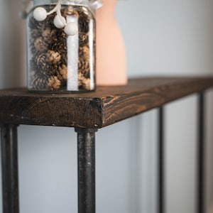 Può includere: Un ripiano in legno marrone scuro con gambe in tubo metallico nero. Il ripiano è vuoto tranne che per un barattolo di vetro pieno di pigne e ornamenti bianchi.